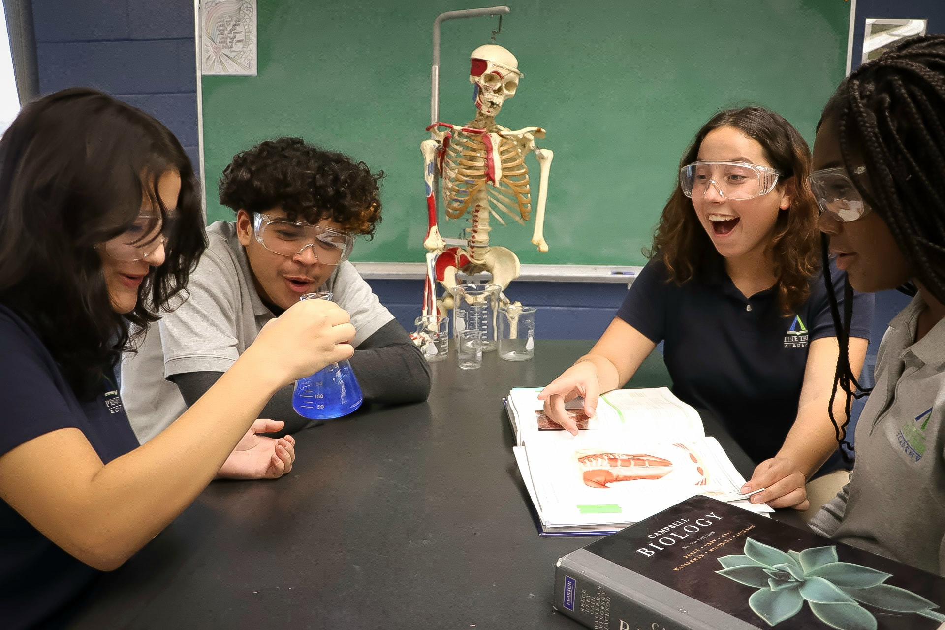 Pine Tree Academy students in safety goggles working with beakers around a lab table in a biology classroom with an anatomical skeleton and chalkboard behind them