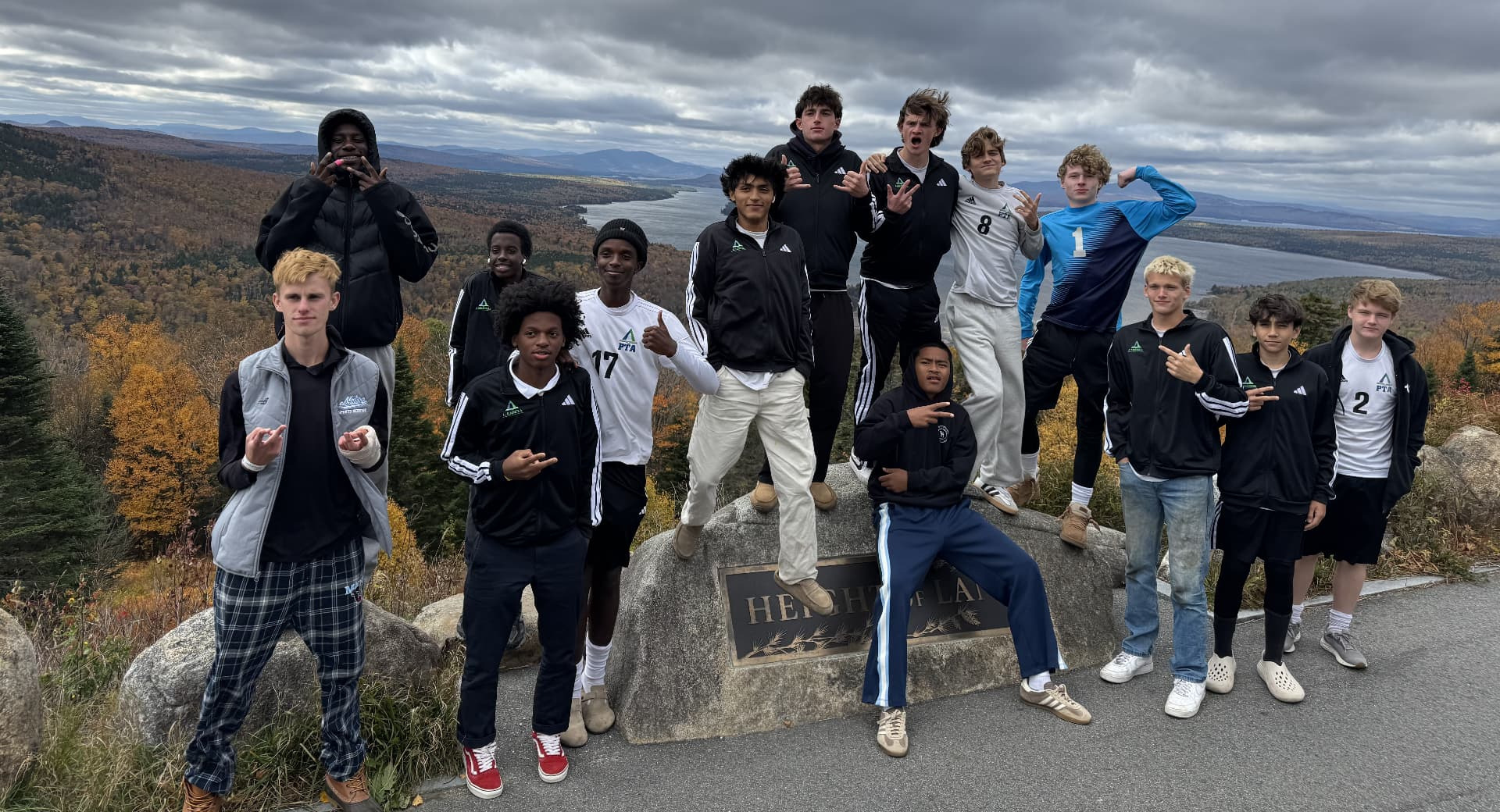 Pine Tree Academy boys varsity soccer team posed on the Height of Land rock in Rangeley, Maine, with Mooselookmeguntic Lake and fall foliage below a dramatic sky