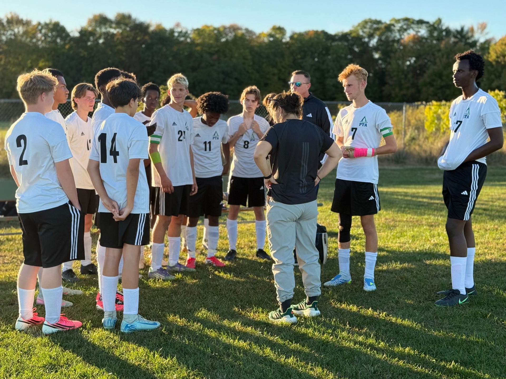 Pine Tree Academy boys varsity soccer players in white jerseys gathered around their coach on the field before the Temple Academy match