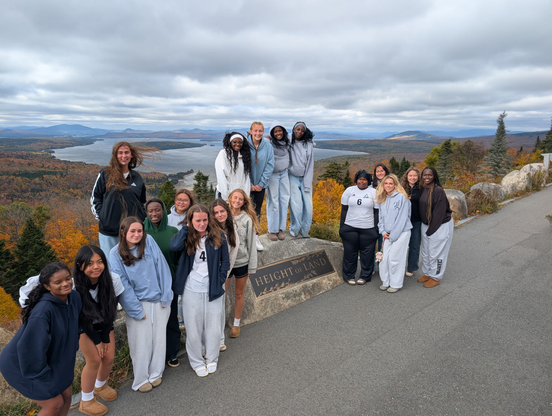 Pine Tree Academy girls varsity soccer team posed together at The Height of Land overlook in Rangeley, Maine, with Mooselookmeguntic Lake and fall foliage in the background